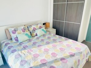 Una cama bien hecha con un edredón de lunares coloridos y almohadas a juego, una mesita de noche con una lámpara, y un ventanal con vistas al exterior.