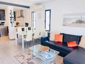 Un espacio habitable moderno con comedor, una mesa de centro de cristal, un sofá negro con cojines naranjas y una cocina al fondo.