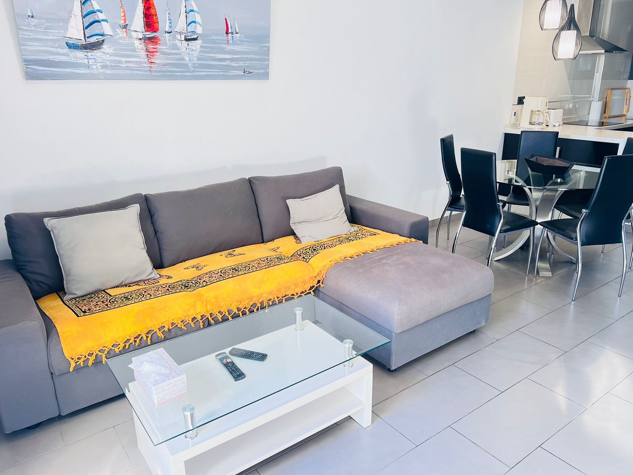 Una sala de estar moderna con un sofá gris, una mesa de centro de vidrio y una manta amarilla. Hay un cuadro de veleros en la pared y un comedor con sillas y una mesa.