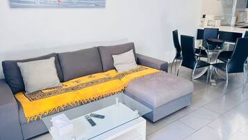 Una sala de estar moderna con un sofá gris, una mesa de centro de vidrio y una manta amarilla. Hay un cuadro de veleros en la pared y un comedor con sillas y una mesa.