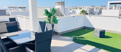 A rooftop terrace with a glass table and chairs, a small artificial lawn, and a view of the city skyline.