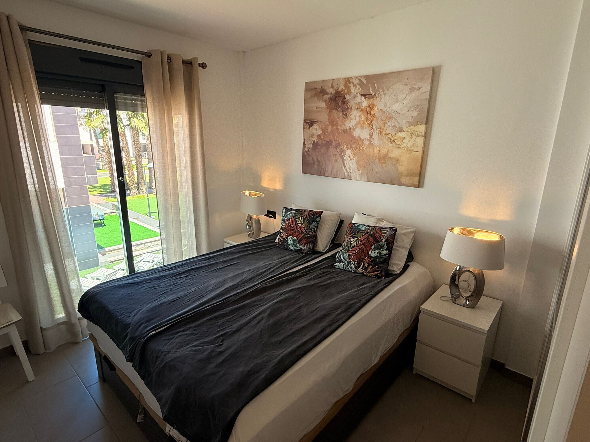 A bedroom with a bed, two bedside lamps, a nightstand, a window with curtains, and a painting on the wall.