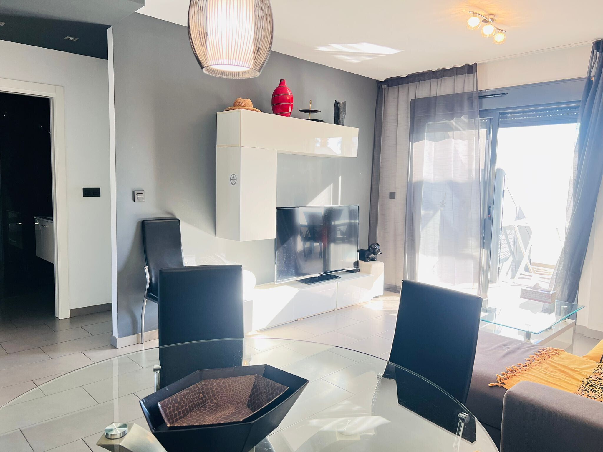 Un salon moderne avec une table basse en verre, des chaises noires, un téléviseur à écran plat et un vase rouge posé sur une console blanche.