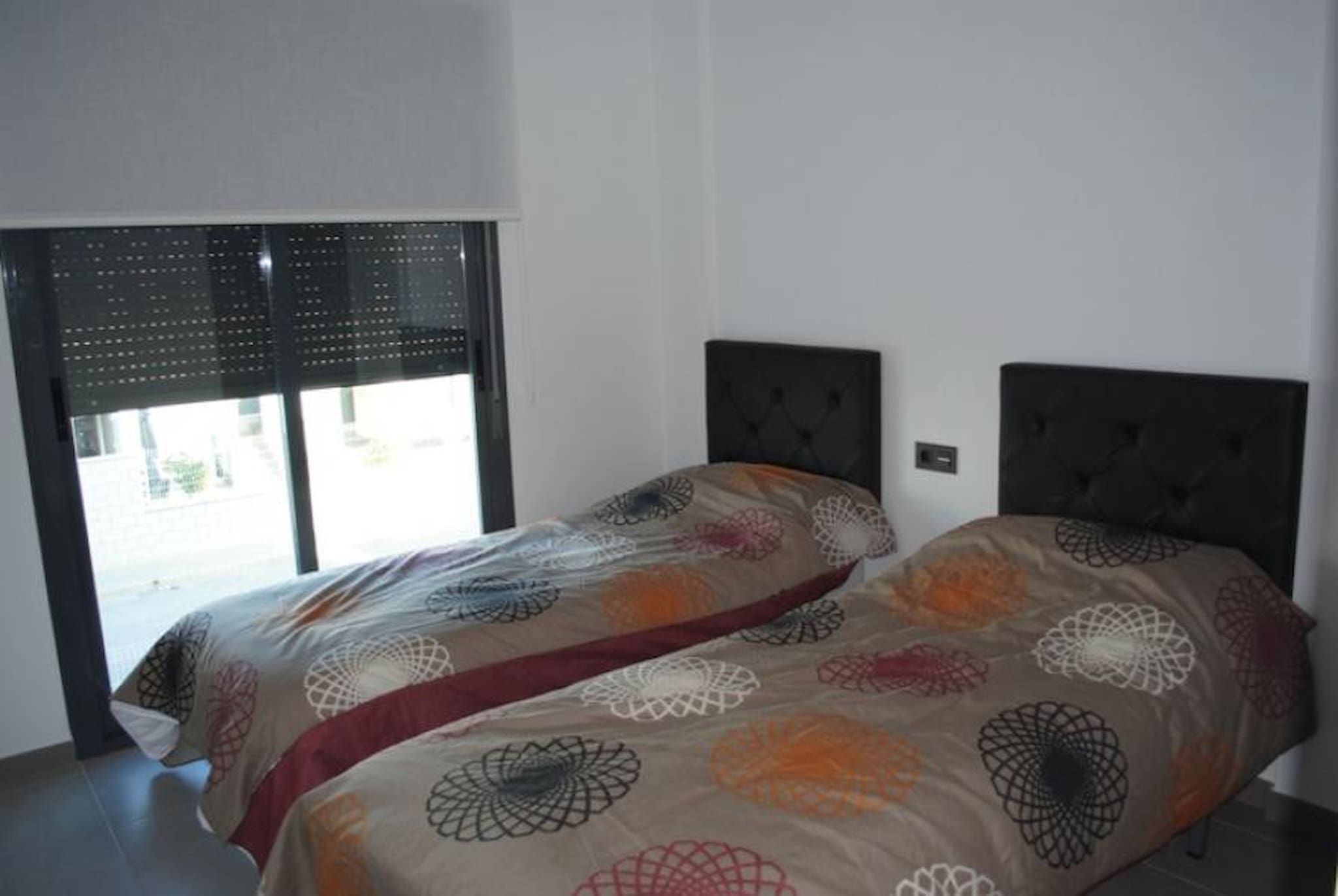 A bedroom with a bed, a window with blinds, and a wall-mounted light switch.