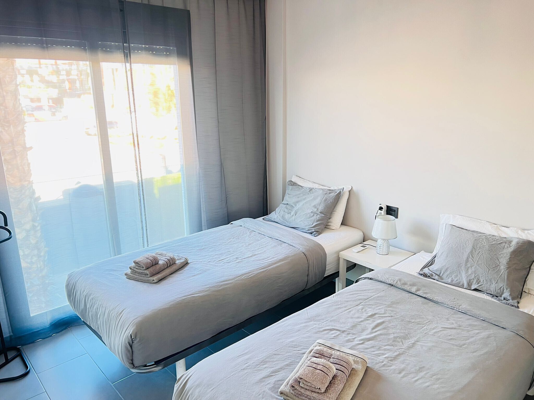 Una habitación de hotel con dos camas individuales, una mesita de noche con lámpara y una ventana con cortinas.