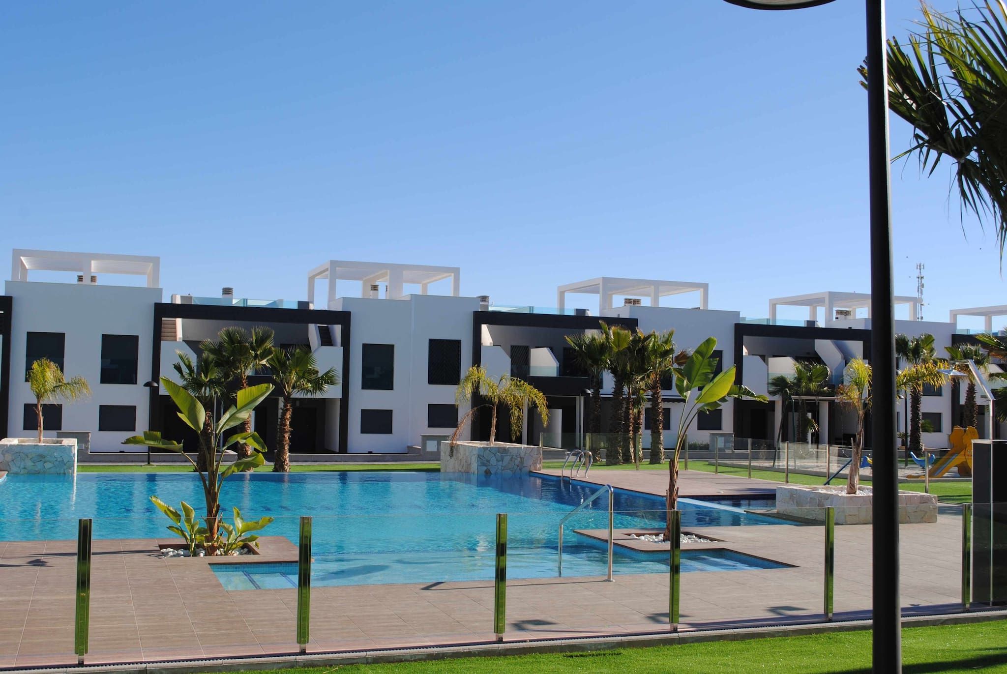 A modern building complex with a central swimming pool, surrounded by palm trees and a clear sky.