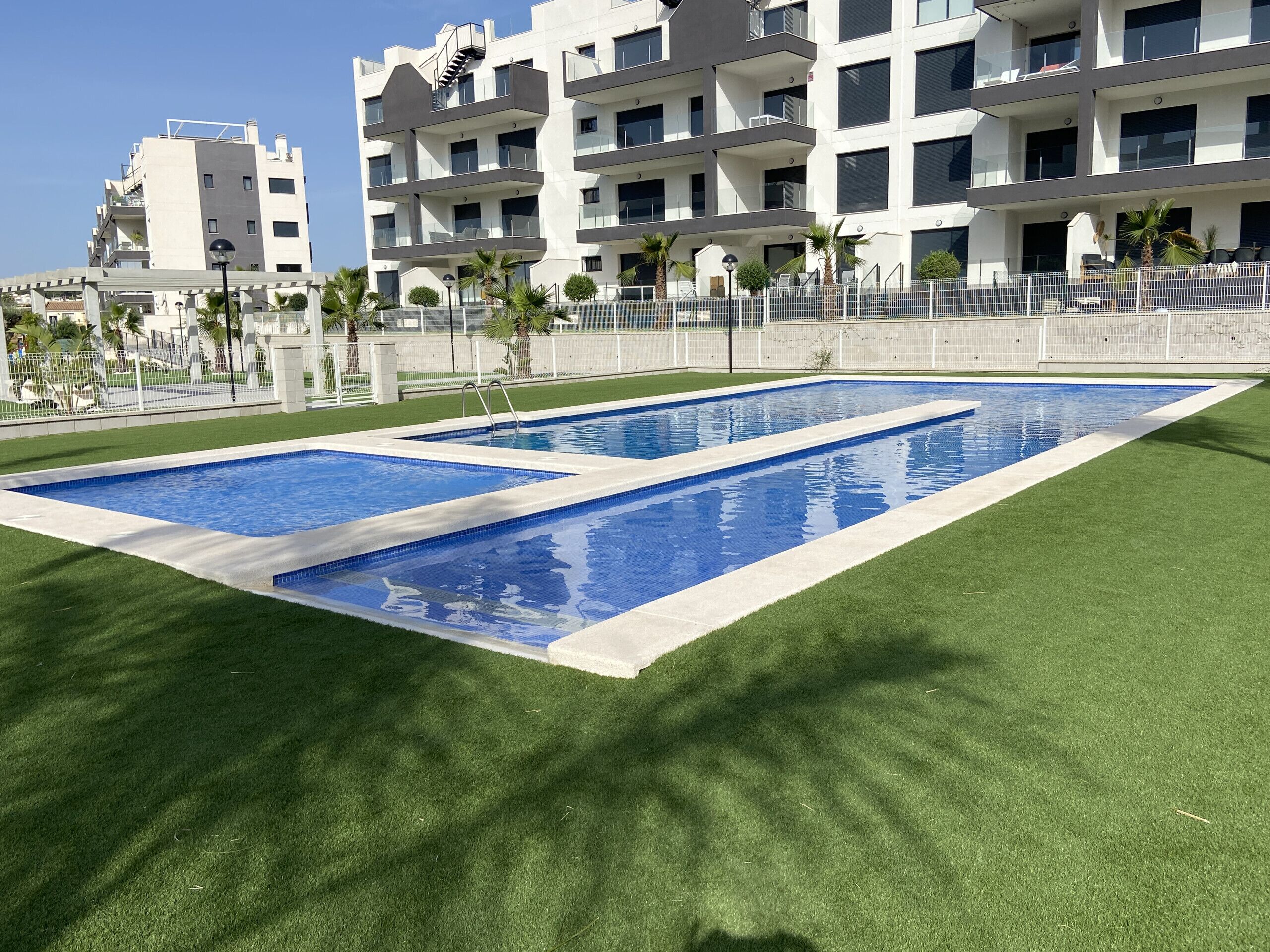 Una piscina rectangular con borde blanco, rodeada de césped artificial, y un edificio moderno con balcones al fondo.