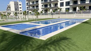 Una piscina rectangular con borde blanco, rodeada de césped artificial, y un edificio moderno con balcones al fondo.