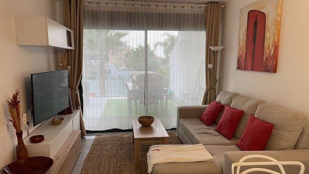 Una sala de estar con sofá, televisión, mesa de centro y vistas a través de una ventana con cortinas.