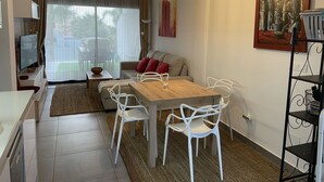 Una sala moderna con una mesa de comedor de madera, sillas blancas, un sofá con cojines rojos, un televisor y un cuadro en la pared.