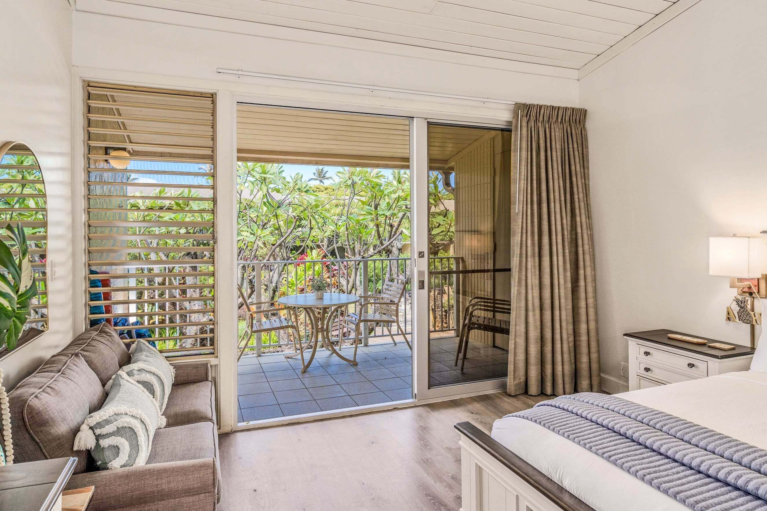 A hotel room with a bed, a sofa, a dining table with chairs, a balcony with a view of greenery, and a sliding glass door.