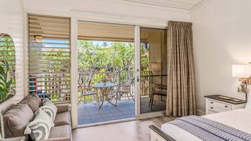 A hotel room with a bed, a sofa, a dining table with chairs, a balcony with a view of greenery, and a sliding glass door.