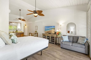 Un dormitorio con una cama, un sofá gris, un escritorio, un ventilador de techo y un cuadro de una playa.