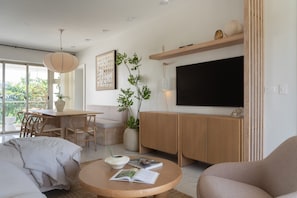 Ein modernes Wohnzimmer mit einer Couch, einem runden Couchtisch mit Büchern, einem Essbereich mit Tisch und Stühlen sowie einem an der Wand befestigten Fernseher.