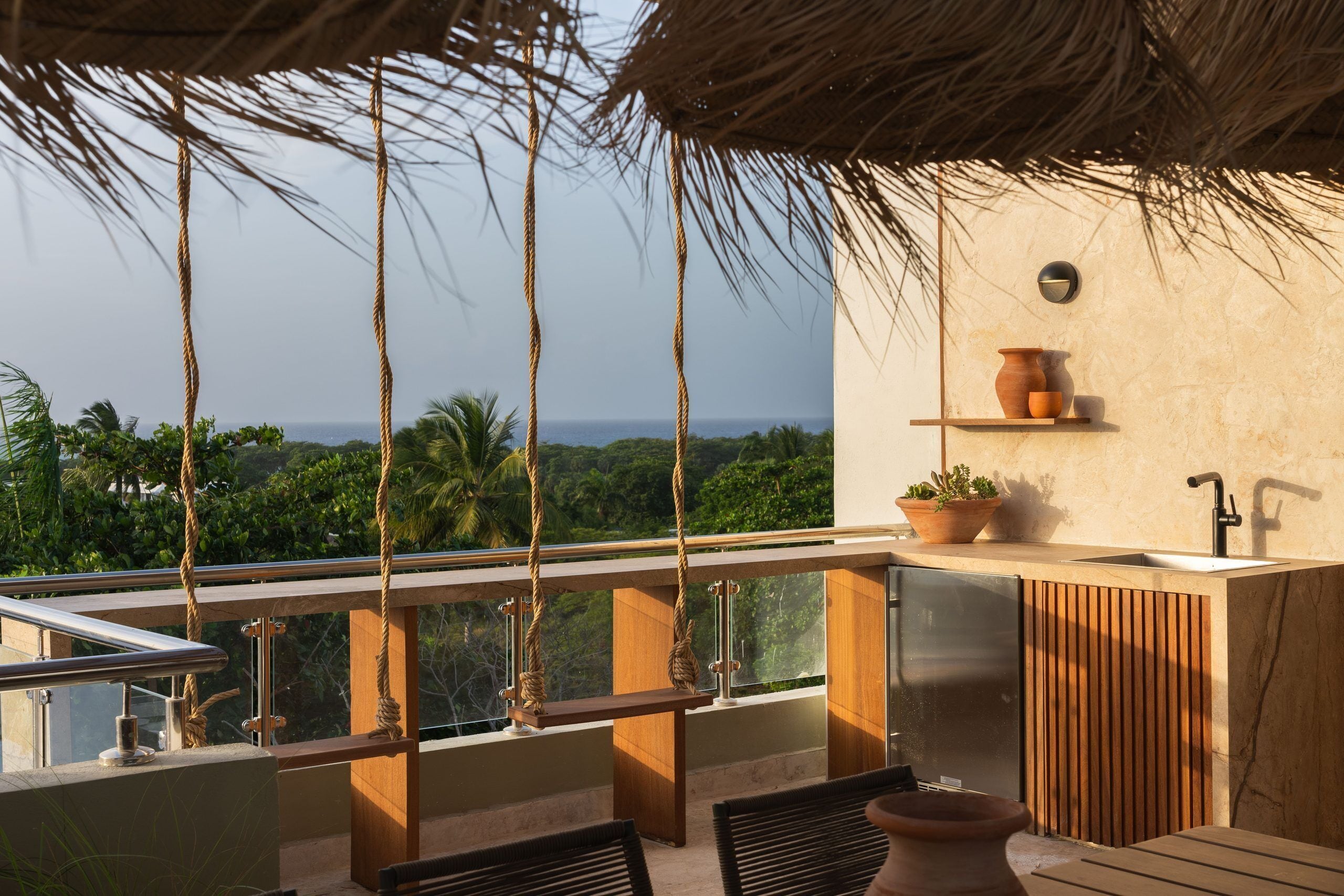 A balcony with a view of the ocean, a wooden table, chairs, a hanging rope, a potted plant, and a wall-mounted shelf with a pot and a cup.