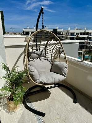 A wicker hanging chair with cushions on a balcony with a view of buildings and a palm tree. - Apartment Playa Flamenca Deluxe (Orihuela)
