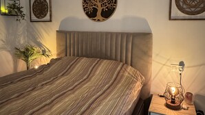 Ein Schlafzimmer mit gestreifter Bettdecke, einem Kopfteil, einem Nachttisch mit Lampe sowie Wanddekorationen, darunter ein Baum und geometrische Muster.
