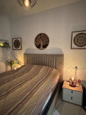 A bedroom with a striped bedspread, a headboard, a bedside table with a lamp, and wall decorations including a tree and geometric patterns. - Apartment Playa Flamenca Deluxe (Orihuela)