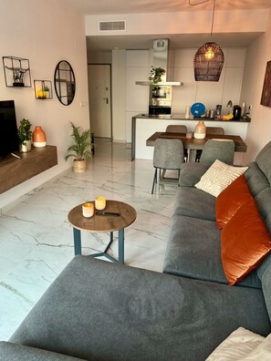A modern living room with a grey sofa, a coffee table, and a dining area with chairs. - Apartment Playa Flamenca Deluxe (Orihuela)