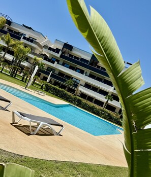 Une piscine entourée de chaises longues, de verdure et d’un bâtiment moderne doté de nombreux balcons.