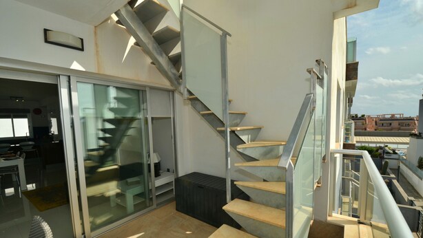 Een modern appartement met een glazen deur naar een balkon, een trap met glazen leuningen en uitzicht op de stad.