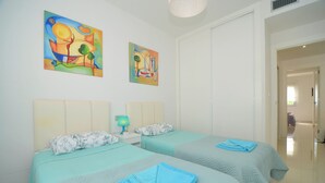 Een hotelkamer met twee bedden, elk voorzien van een blauw dekbed en kussens, een nachtkastje met een lamp, en twee kleurrijke schilderijen aan de muur.
