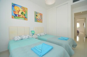 Ein Hotelzimmer mit zwei Betten, jeweils mit blauer Bettdecke und Kissen, einem Nachttisch mit Lampe sowie zwei farbenfrohen Bildern an der Wand.