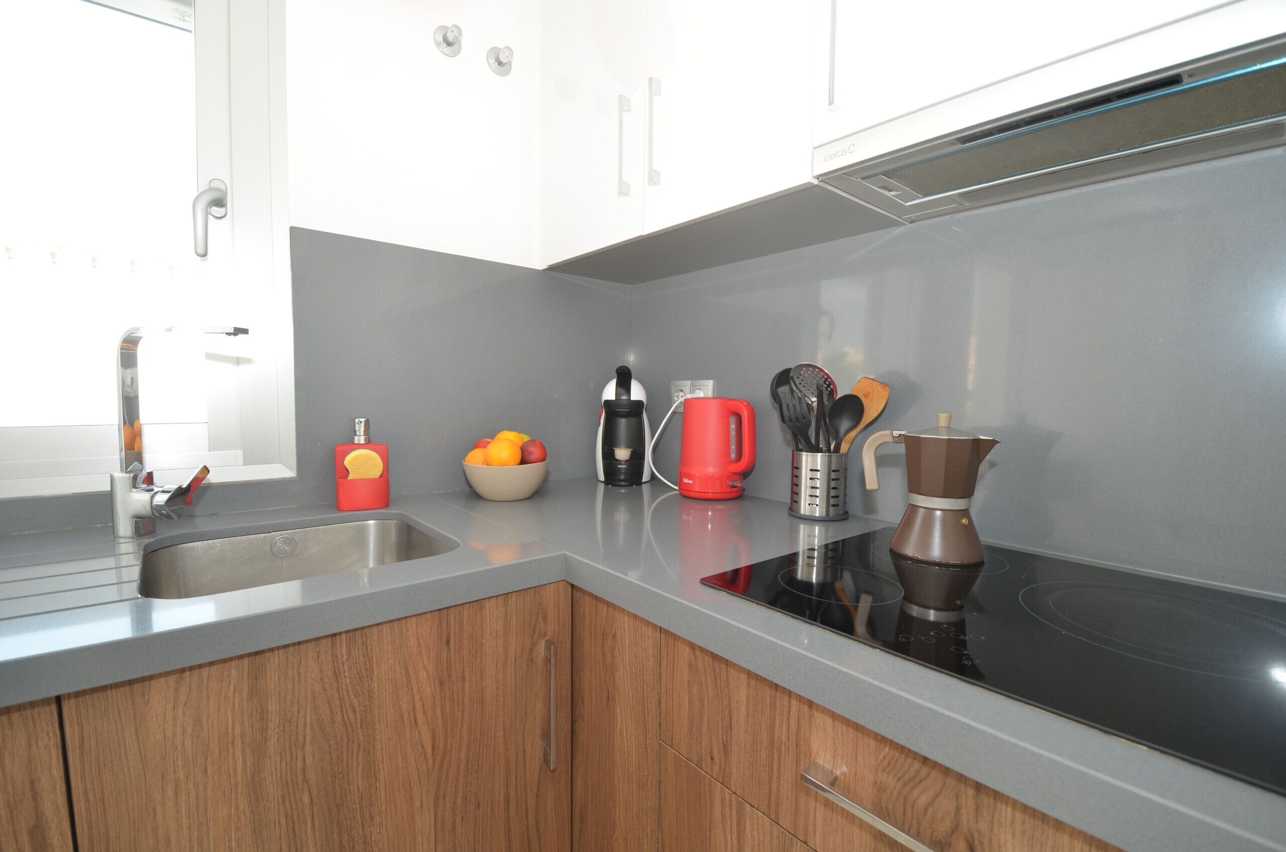 Una cucina moderna con lavello in acciaio inossidabile, bollitore rosso, macchina da caffè e una ciotola di frutta.