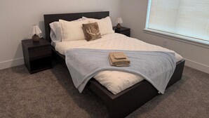 A neatly made bed with a light-colored bedspread and pillows, a folded towel on the bed, and a window with blinds to the right.