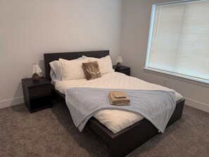 A neatly made bed with a light-colored bedspread and pillows, a folded towel on the bed, and a window with blinds to the right.