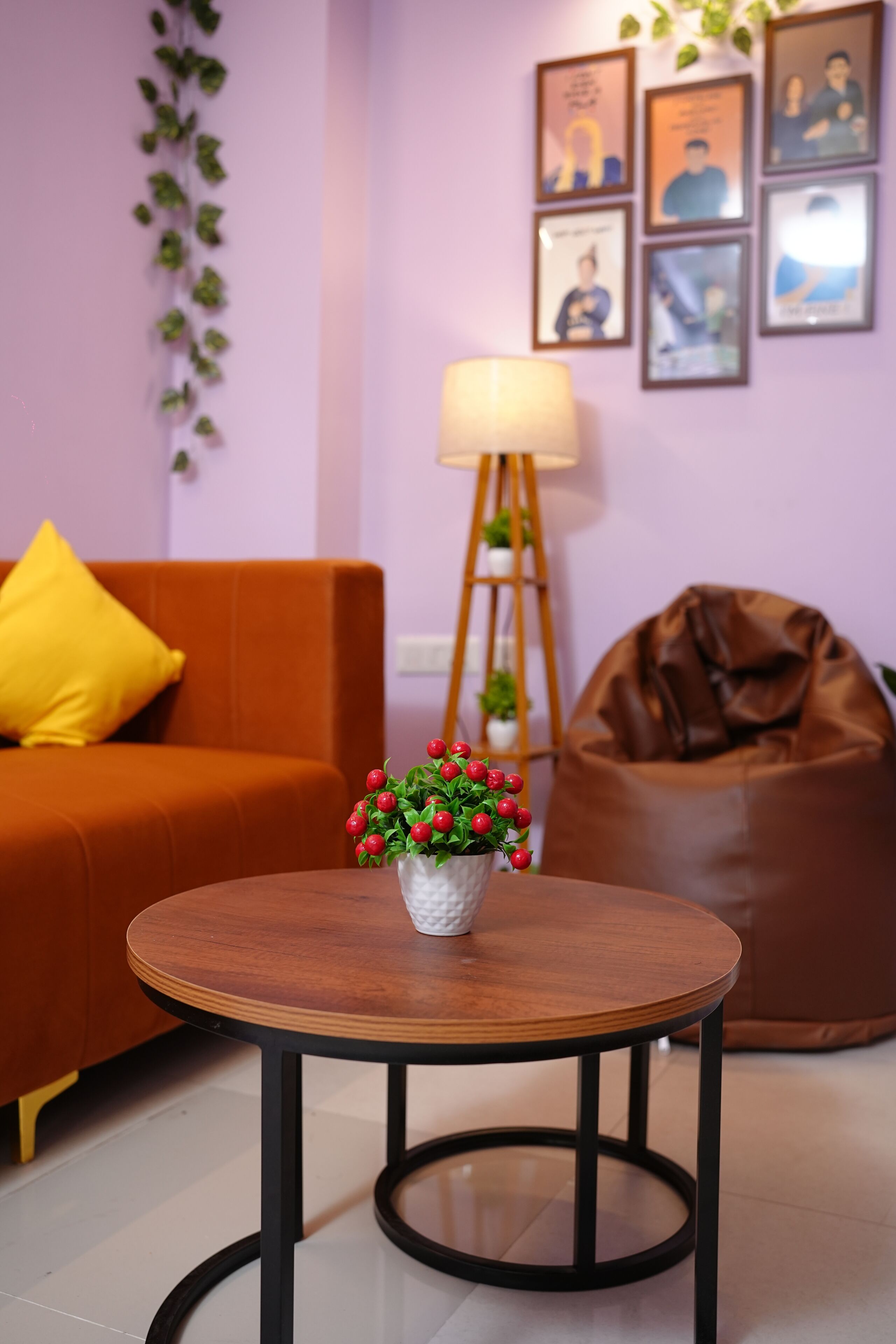 A living room with a round wooden table, a sofa with a yellow pillow, a brown bean bag, a floor lamp, and framed pictures on the wall.