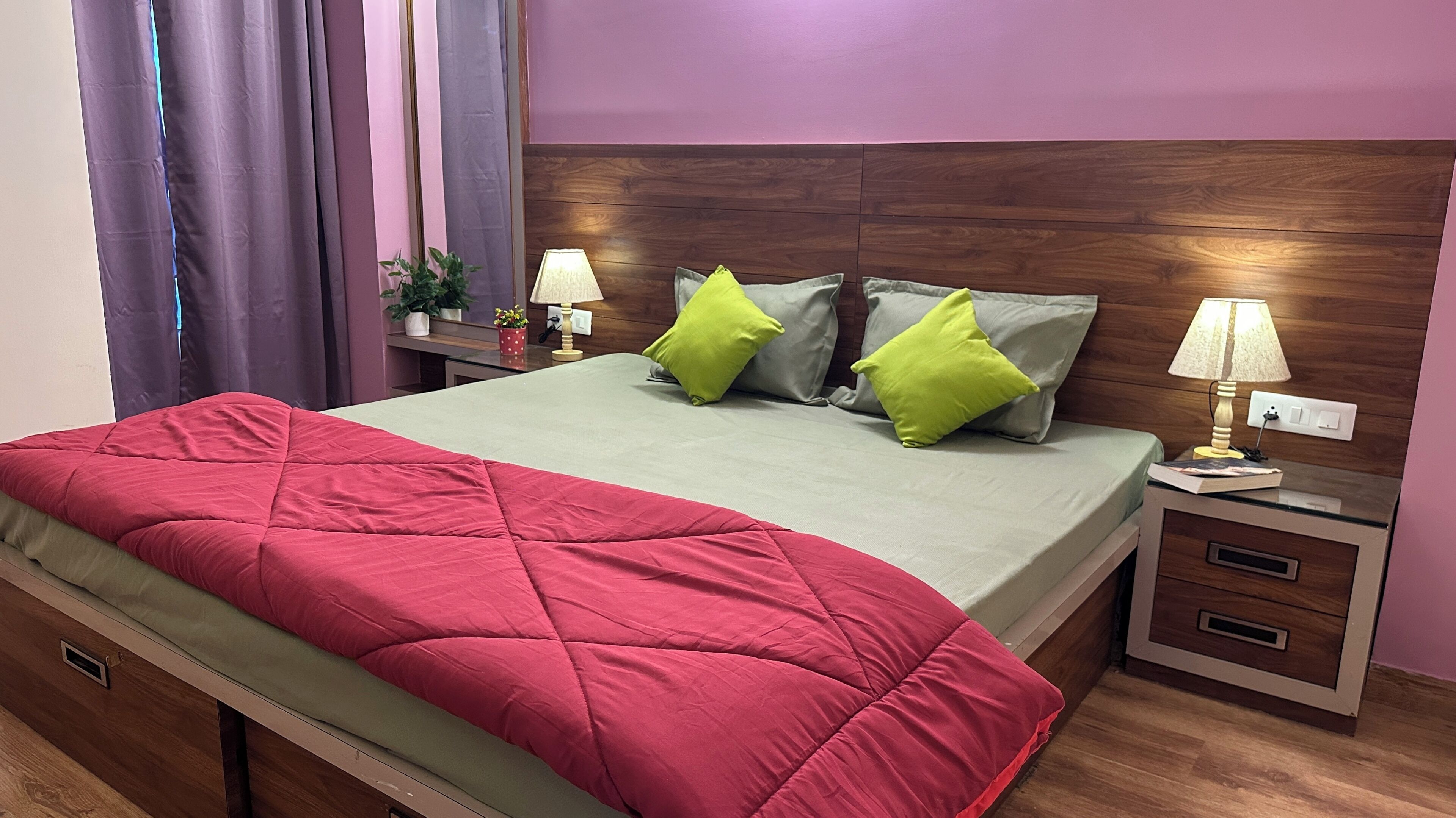 A neatly made bed with a red and green quilt, two bedside lamps, and a wooden nightstand with a drawer.