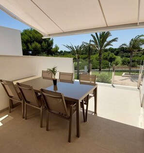 A covered outdoor dining area with a table and chairs, a potted plant, and a view of greenery and trees. - Penthouse Romerogolf (Pilar de la Horadada)