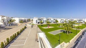 A modern urban street with white buildings, a green park area with palm trees, and a playground. - Penthouse Oasis Beach IX Punta Prima (Torrevieja)
