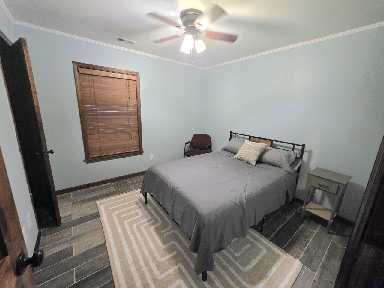A bedroom with a bed, a chair, a bedside table, a ceiling fan, and a window with blinds.