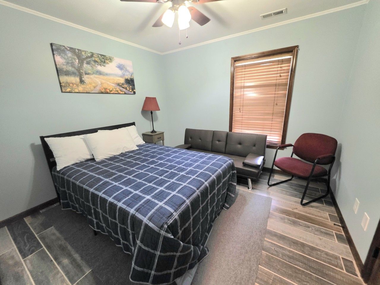 A bedroom with a bed, a chair, a nightstand, a lamp, and a window with blinds.