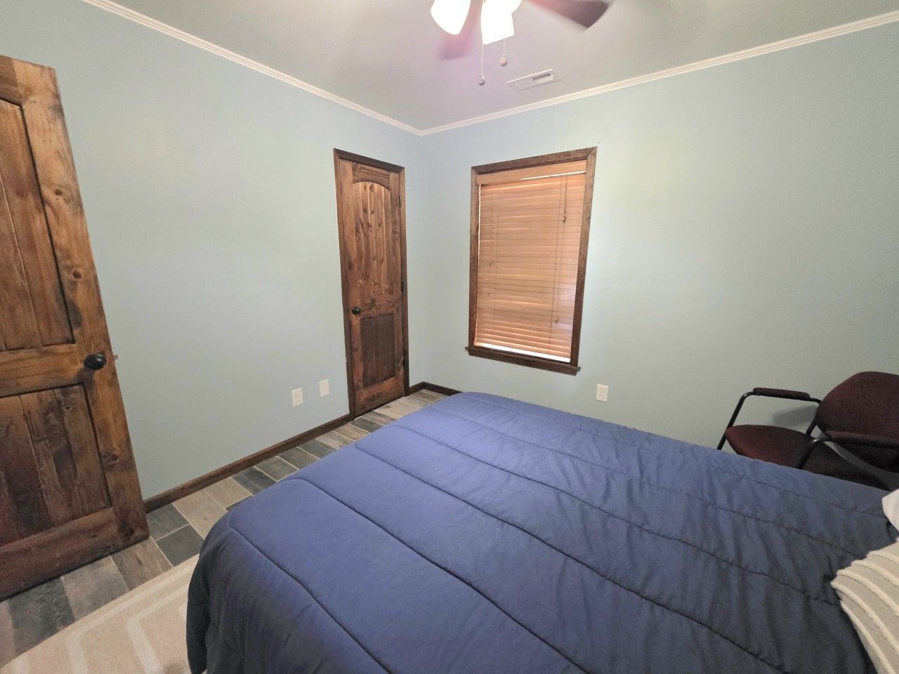 A bedroom with a bed, two doors, a window with blinds, a chair, and a ceiling fan.