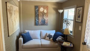 A living room with a beige sofa, a map on the wall, a painting, a lamp, and a window with a windmill decoration. - Desert Rose Located Near Texas Tech (Lubbock)