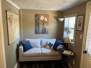 Ein Wohnzimmer mit einem beige farbenen Sofa, einer Karte an der Wand, einem Gemälde, einer Lampe und einem Fenster mit Windmühlenverzierung.
