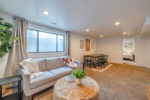 A living room with a sofa, a dining table with chairs, a coffee table with a plant, and a large window with curtains. - Cozy Stay | Patio | Full Kitchen | Long Term Ready (Sandy)