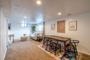 A room with a wooden dining table, four chairs, a sofa, a TV, and artwork on the walls. - Cozy Stay | Patio | Full Kitchen | Long Term Ready (Sandy)