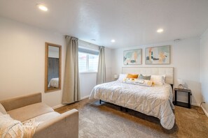 A bedroom with a bed, a chair, a nightstand, a lamp, and artwork on the wall. - Cozy Stay | Patio | Full Kitchen | Long Term Ready (Sandy)