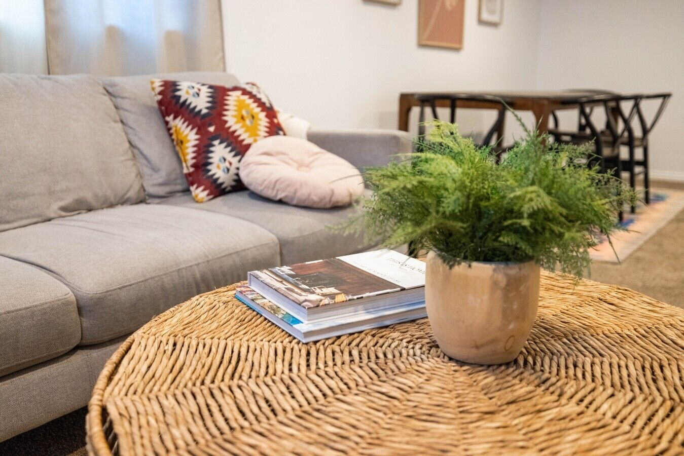 Un saló amb un sofà gris, un coixí decoratiu estampat, una taula de centre trenada amb una planta en test i una pila de revistes.