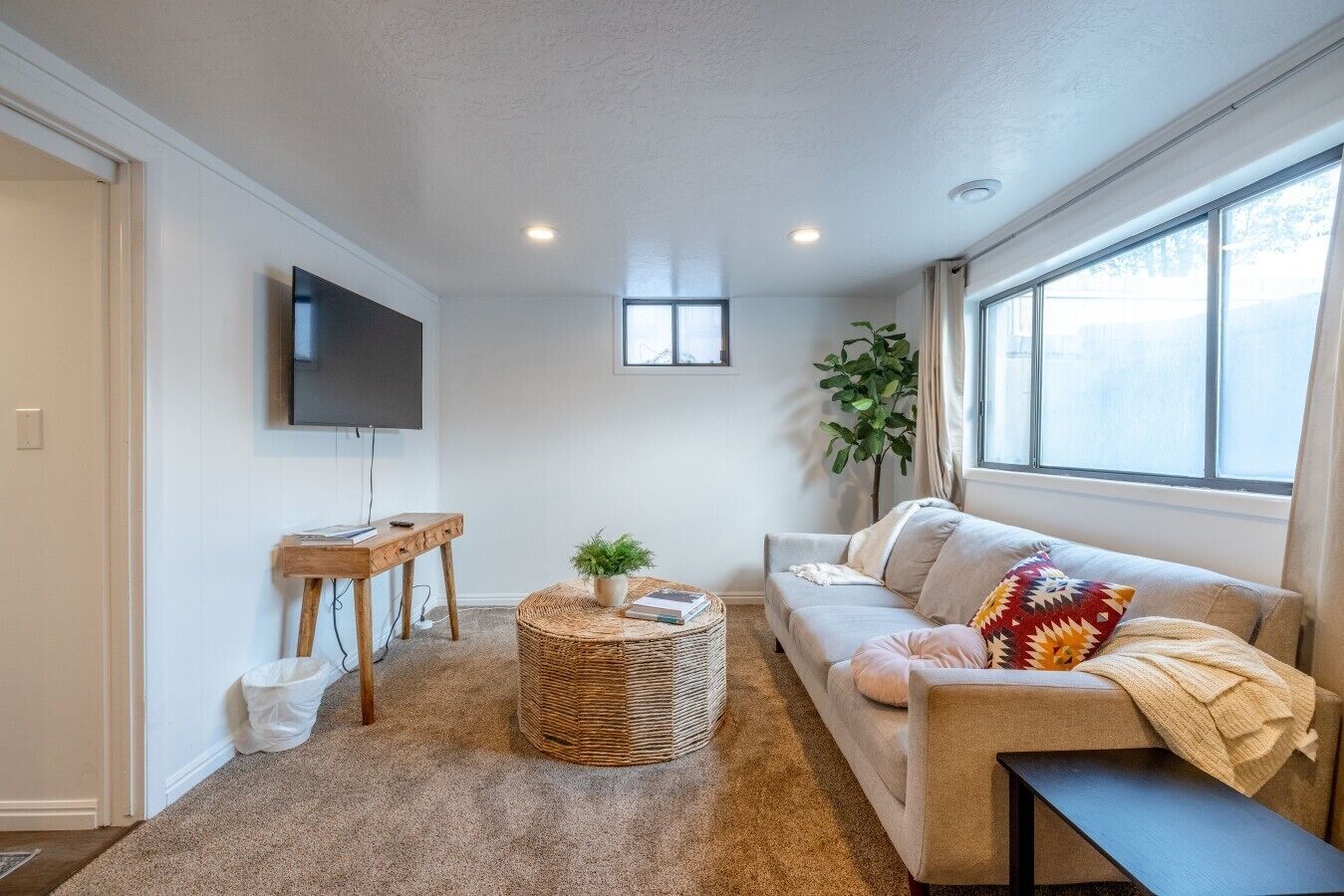 A living room with a sofa, a coffee table, a TV mounted on the wall, and a window providing natural light.