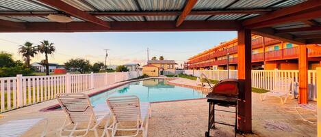 A covered swimming pool with lounge chairs, a barbecue grill, and a multi-story building in the background.