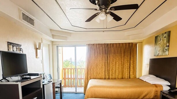 A hotel room with a bed, a desk with a television, a ceiling fan, and a view of the outdoors. - Oro North Beach Inn (Corpus Christi)
