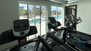 Et fitnessrum med løbebånd, en motionscykel og en romaskine. Der er et stort vindue med udsigt til en swimmingpool og udendørs siddepladser.