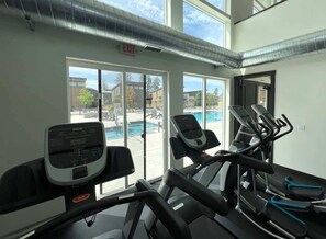 Ein Fitnessraum mit Laufbändern, einem Ergometer und einem Rudergerät. Ein großes Fenster bietet Blick auf einen Pool und Sitzgelegenheiten im Freien.