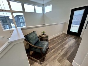 A room with a wooden floor, a blue chair, a small table with a plant, and a large window with a view of buildings. - 40 Mins to Glacier Park, 5 Mins to City Beach & DT (Whitefish)