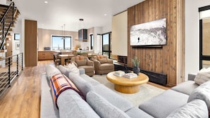 A modern living room with a large flat-screen TV, a fireplace, and a staircase leading to an upper level. - Luxury Ski-In&Out | Private Spa | Mountain Views (Whitefish)